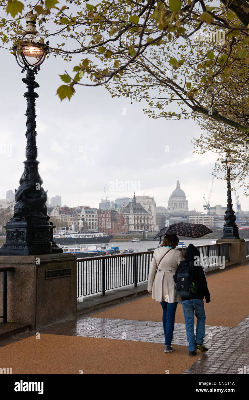 Due persone che camminano con ombrello sul Queens a piedi South Bank in una molla doccia illuminata con luce in stile vittoriano e la Cattedrale di St Paul Foto Stock