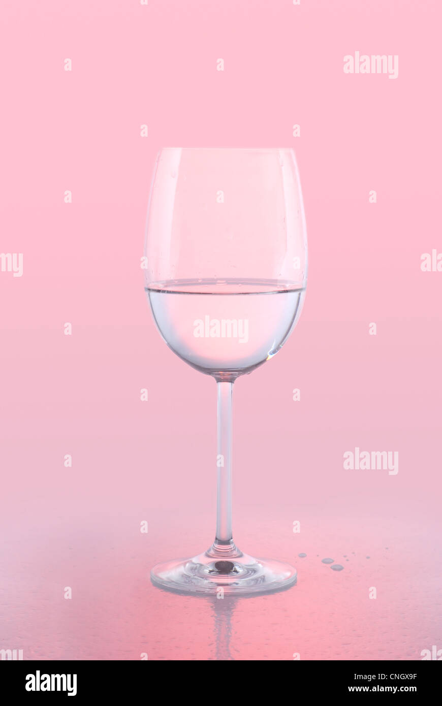 Un cristallo vino bianco vetro acqua tenuta contro un sfondo rosa con la riflessione e gocce d'acqua. Foto Stock