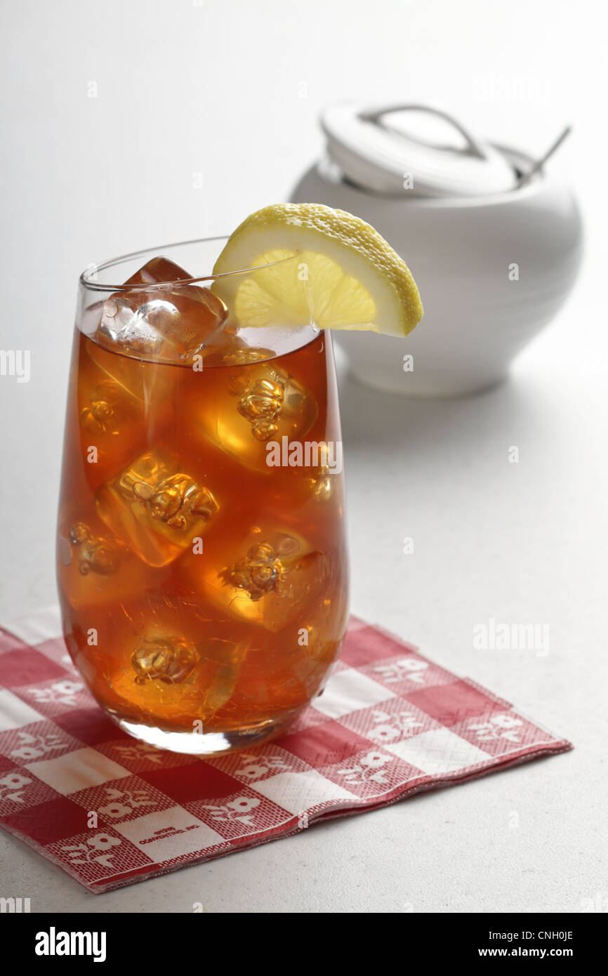 Un bicchiere di tè freddo al limone su un rosso/bianco igienico, con cubetti di ghiaccio e una tazza di zucchero sul lato destro. Foto Stock