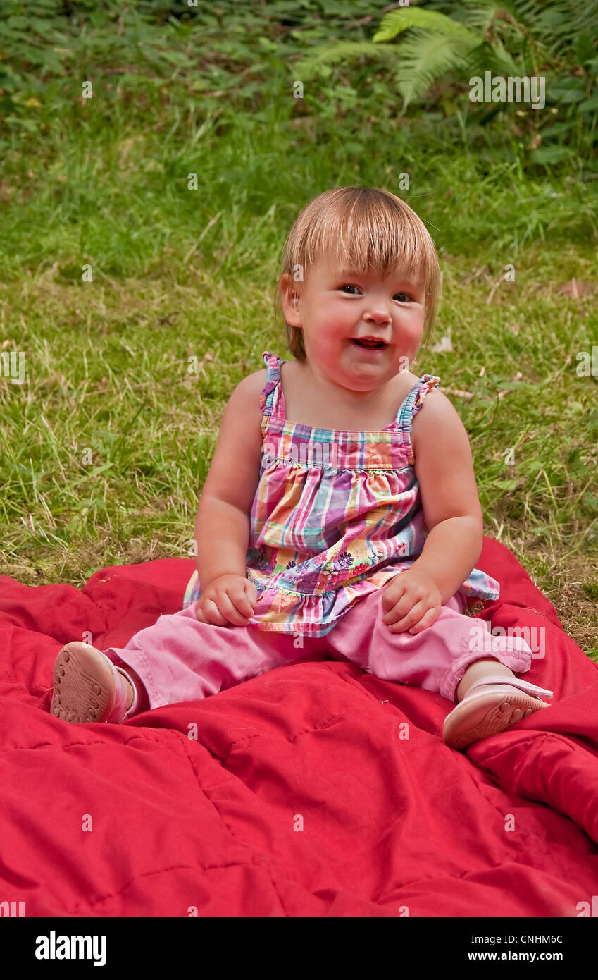 Questo grazioso felice anno vecchio toddler girl, è seduto fuori su una coperta di rosso, sorridente e felice. Sta indossando un plaid shirt. Foto Stock