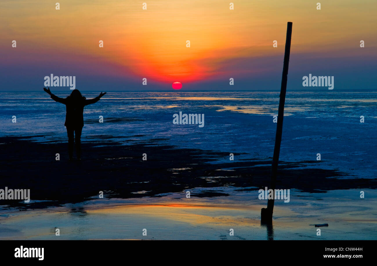 Silhouette di una donna in piedi presso l'estuario del Mare del Nord a marea di declino meditando con le braccia sollevate davanti al tramonto, Germania, Bassa Sassonia, Cuxhaven Foto Stock