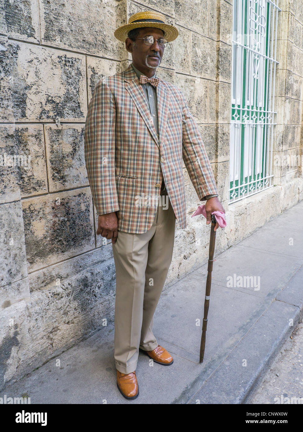 Un buio anziano gentiluomo cubano in un diportista più paglia hat, controllato tuta e bow tie sorge da un muro di pietra a l'Avana. Foto Stock