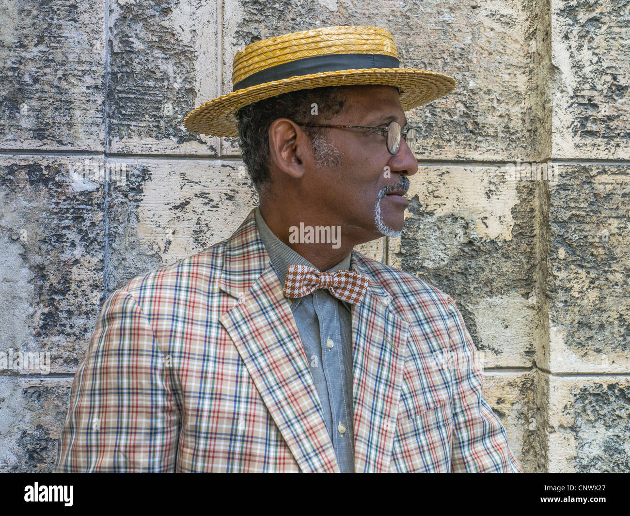 Un buio anziano gentiluomo cubano in un diportista più paglia hat, controllato tuta e bow tie sorge da un muro di pietra a l'Avana. Foto Stock