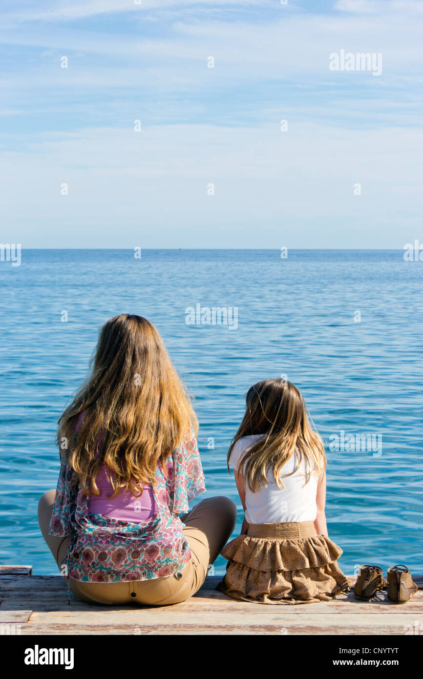 Giovane donna e bambina sat sulla fine del pontile in legno che guarda al mare. Foto Stock