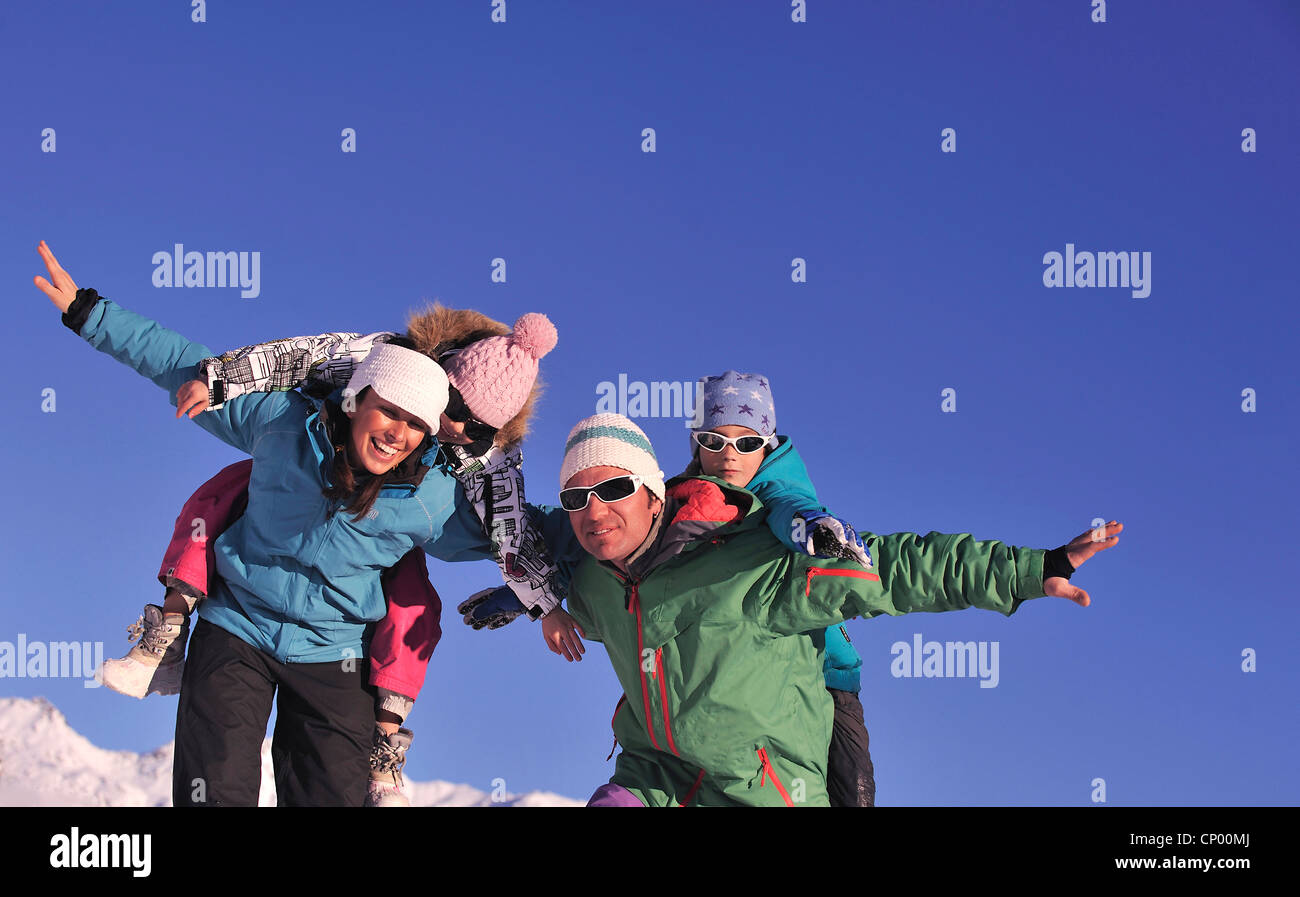 Il padre e la madre in piedi su un snowbound pendio di montagna che trasportano i loro due figli piggyback, Francia Foto Stock