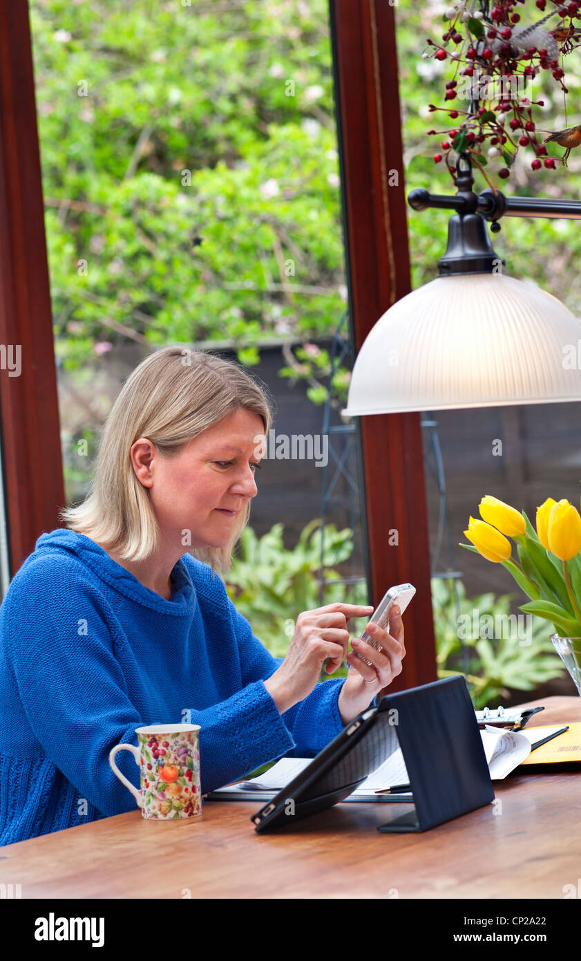 TABLET e SMARTPHONE WIFI A BANDA LARGA Bionda donna che lavora a casa in giardino d'inverno utilizzando il suo Apple iPad tablet computer e smartphone iPhone Foto Stock