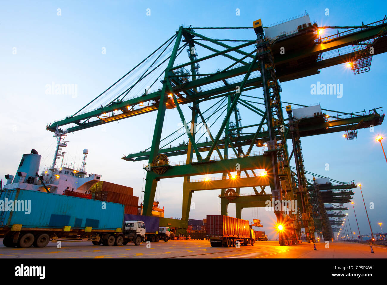 Gru, container e camion a un porto di spedizione durante il crepuscolo Foto Stock