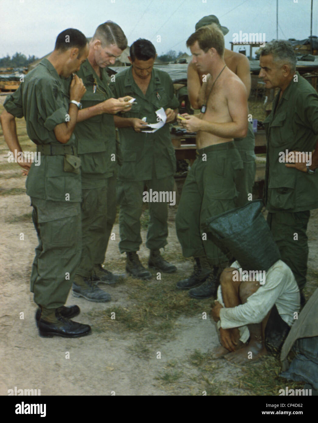 Guerra del Vietnam. Un sospetto Viet Cong siede sulla terra con un sacco nero sopra la sua testa mentre diversi soldati USA esaminare documenti. Foto Stock