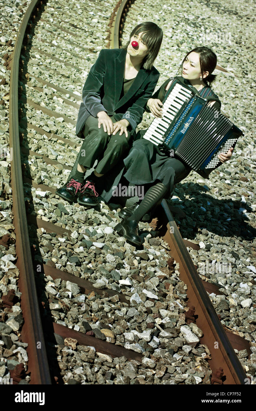 Un triste clown giovane seduto sui binari della ferrovia - la donna suona la fisarmonica Foto Stock