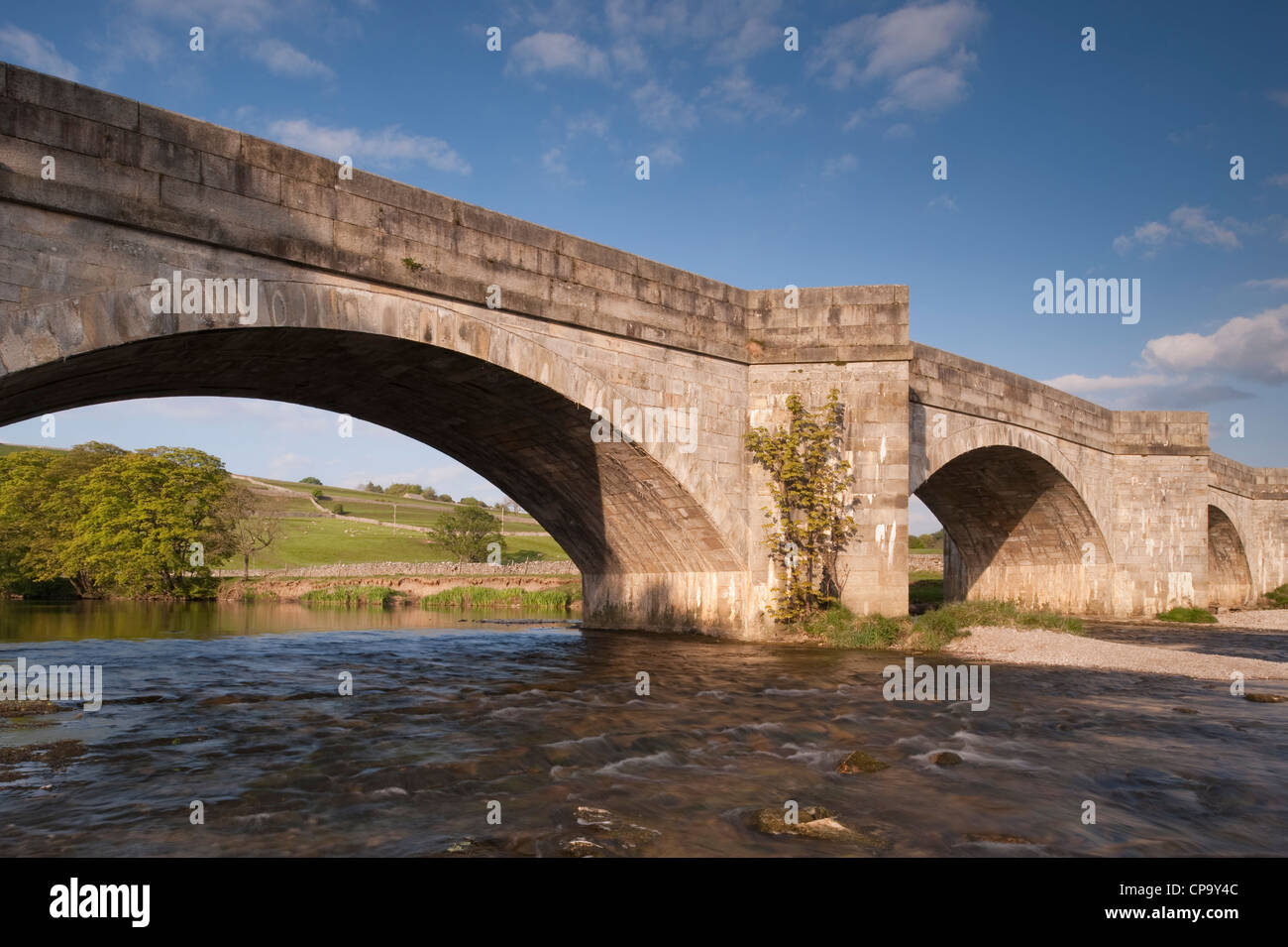 Soleggiato e panoramico, rurale vista del fiume del centro storico, pietra, ponte arcuato spanning che scorre acqua di fiume Wharfe - Burnsall, Yorkshire Dales, Inghilterra, Regno Unito. Foto Stock