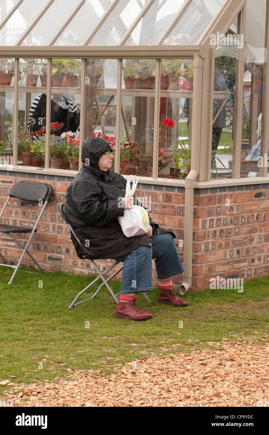 Seduta in pioggia (impermeabile cappuccio) signora è wet & miserabile sulla giornata fuori (da stand commerciali serra) - RHS Tatton Park flower show, Cheshire, Regno Unito. Foto Stock