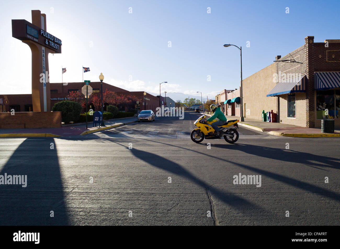 Un motociclo giallo rigidi mediante il Pueblo Bank & Trust segno che getta una lunga ombra. Centro storico cittadino, Salida, CO Foto Stock