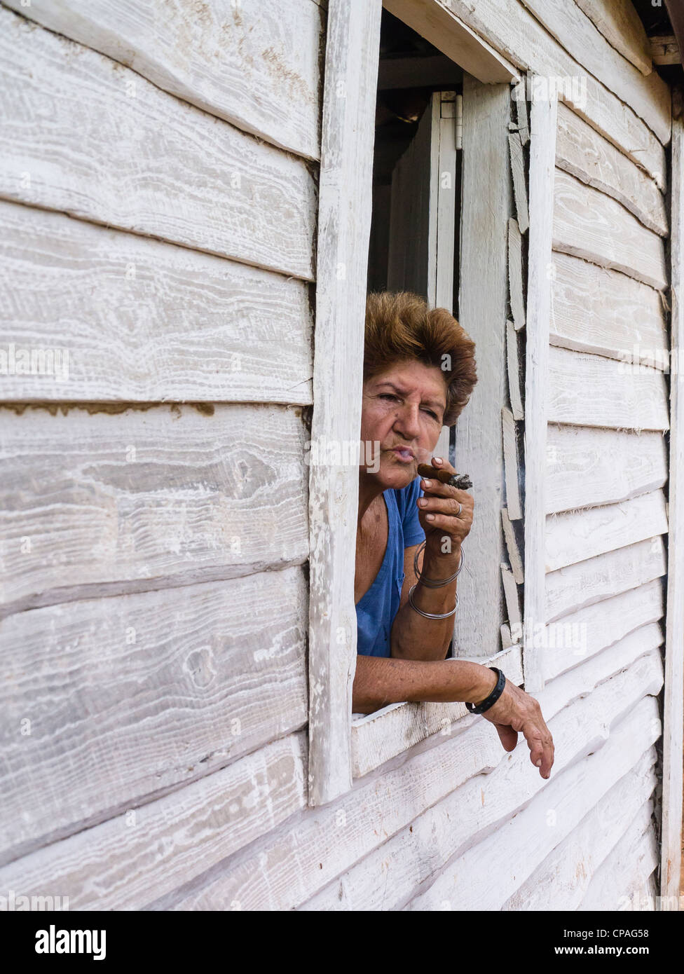 Un cubano femmina senior citizen si inclina fuori da una finestra aperta della sua facciata in legno casa di fumare un sigaro in Viñales, Cuba. Foto Stock