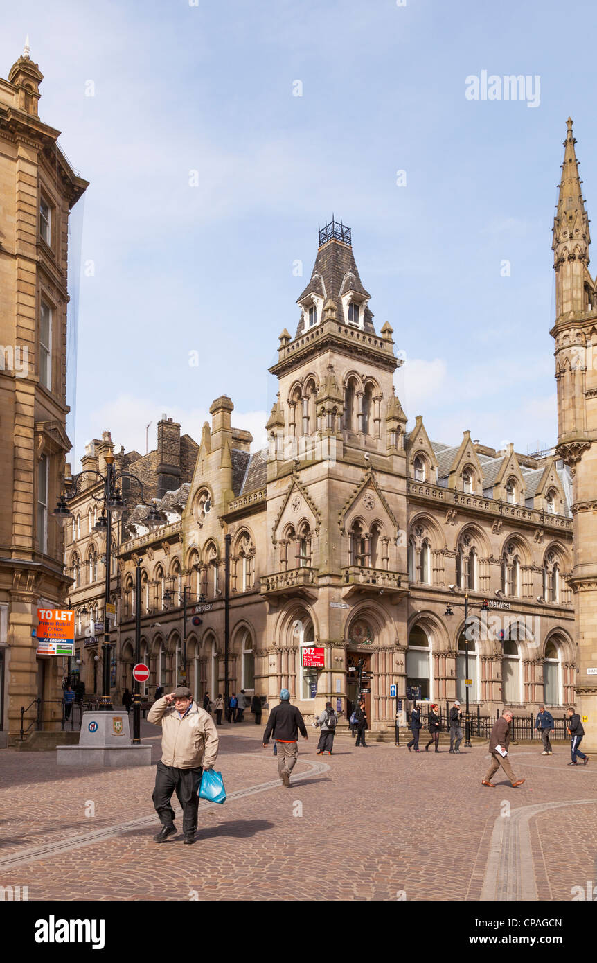 Un vecchio uomo in un tappo piatto cammina davanti a vecchi edifici in pietra arenaria a Bradford, West Yorkshire, Inghilterra, Foto Stock