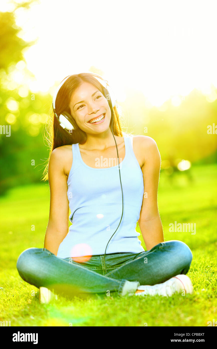 Bella felice multirazziale caucasico Cinese / Asian giovane donna seduta al sole ascoltando la musica sulle cuffie in posizione di parcheggio Foto Stock