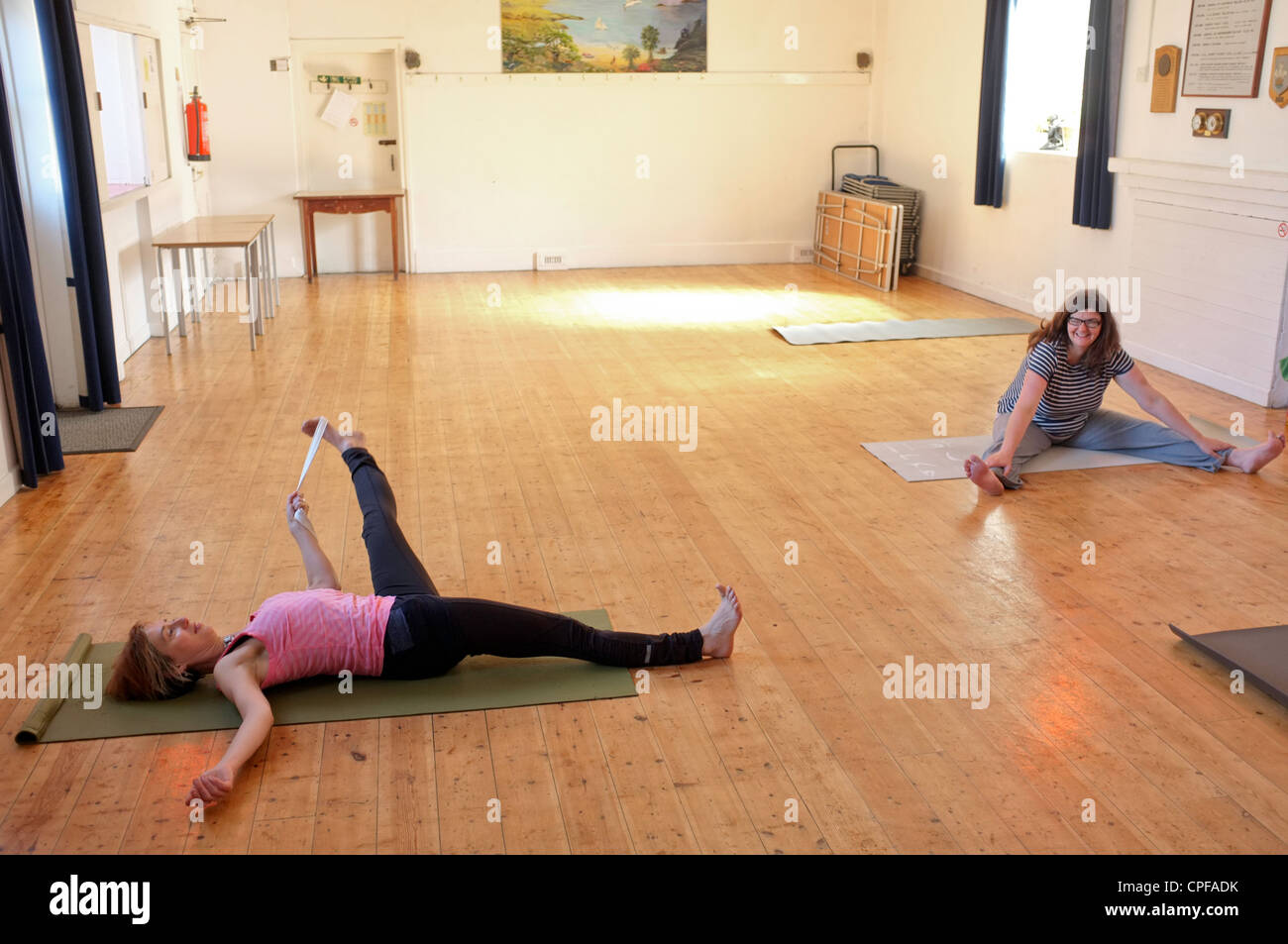 Due donne il riscaldamento prima di una lezione di yoga tenuto in un villaggio hall in Cornwall, Regno Unito Foto Stock