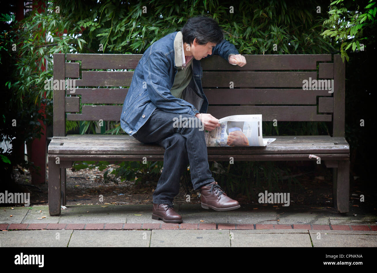 Uomo su una panchina lettura giornali stranieri in Manchester Regno Unito a Chinatown Foto Stock