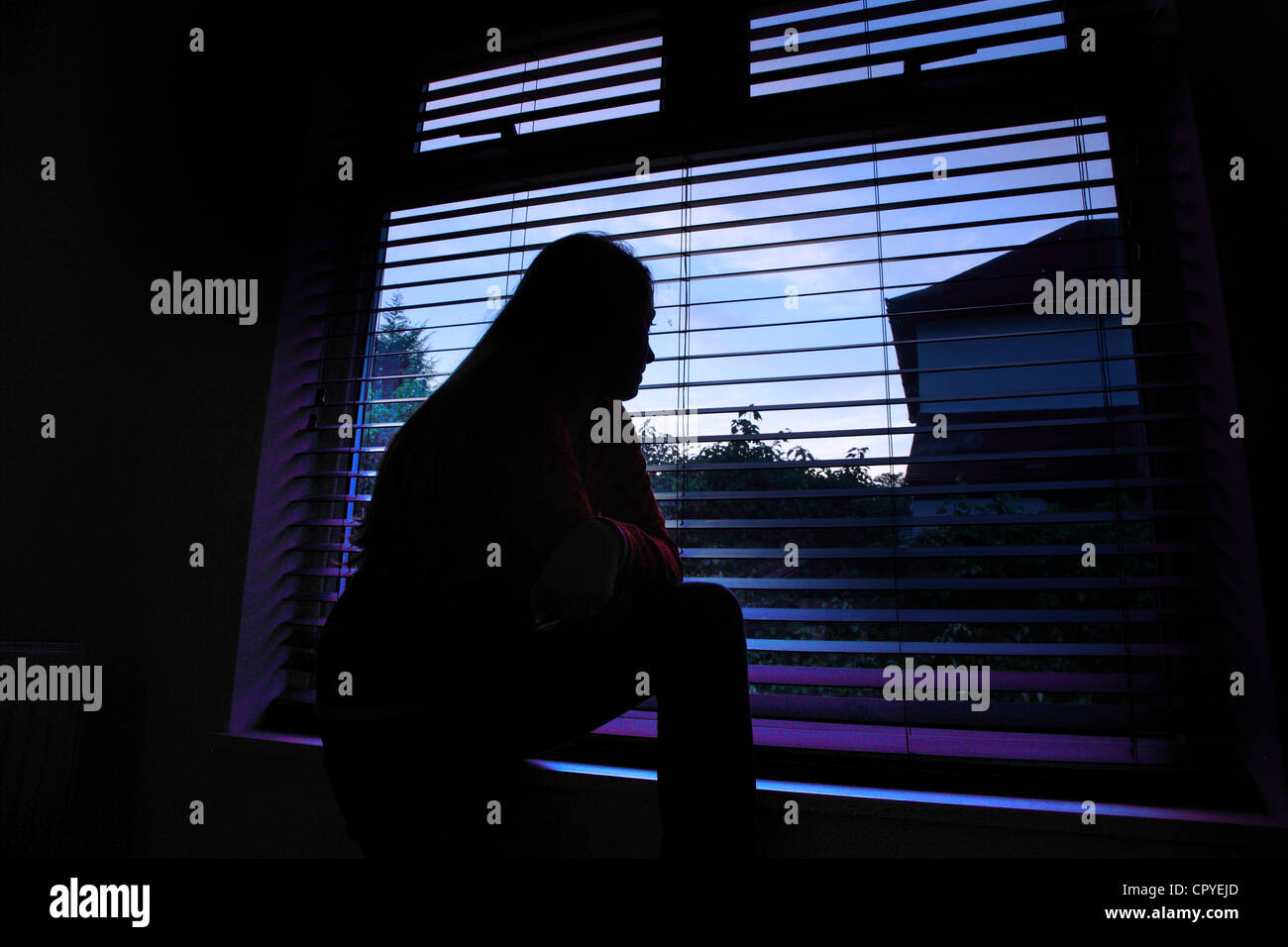 Giovane donna seduta da sola in una stanza buia guardando fuori attraverso una finestra cieca. Foto Stock