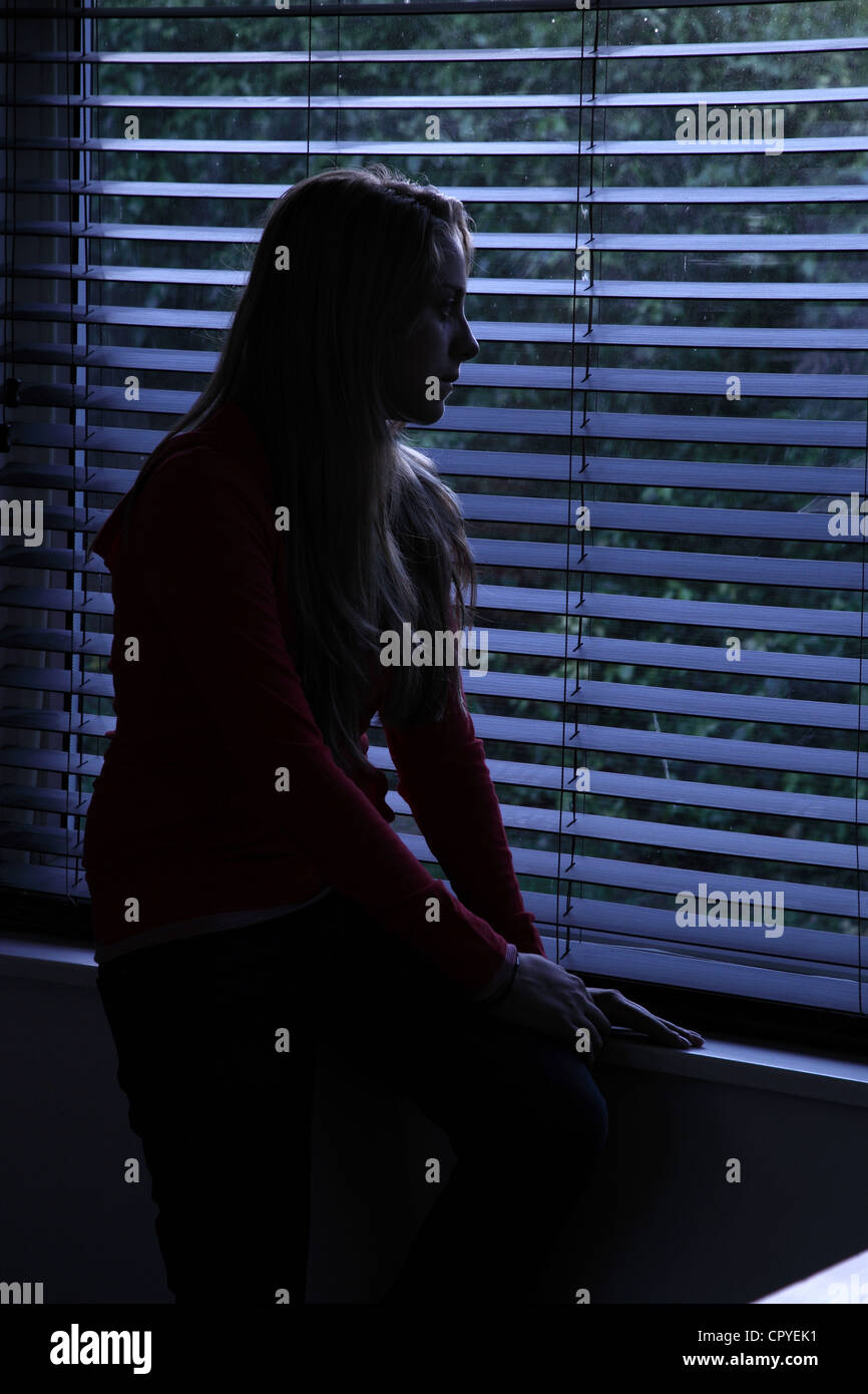 Giovane donna seduta da sola in una stanza buia guardando fuori attraverso una finestra cieca. Modello di proprietà/rilasciato. Foto Stock