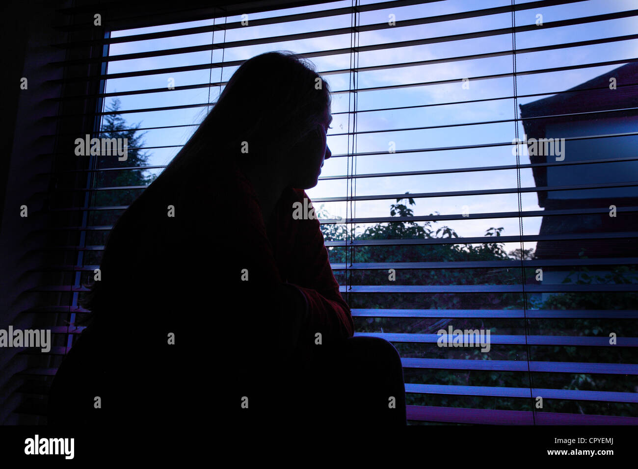 Giovane donna seduta da sola in una stanza buia guardando fuori attraverso una finestra cieca. Foto Stock