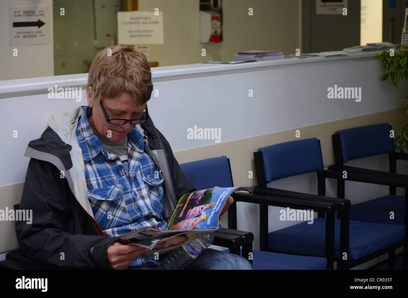 Donna seduta in un ospedale in sala d'attesa leggendo una rivista Foto Stock