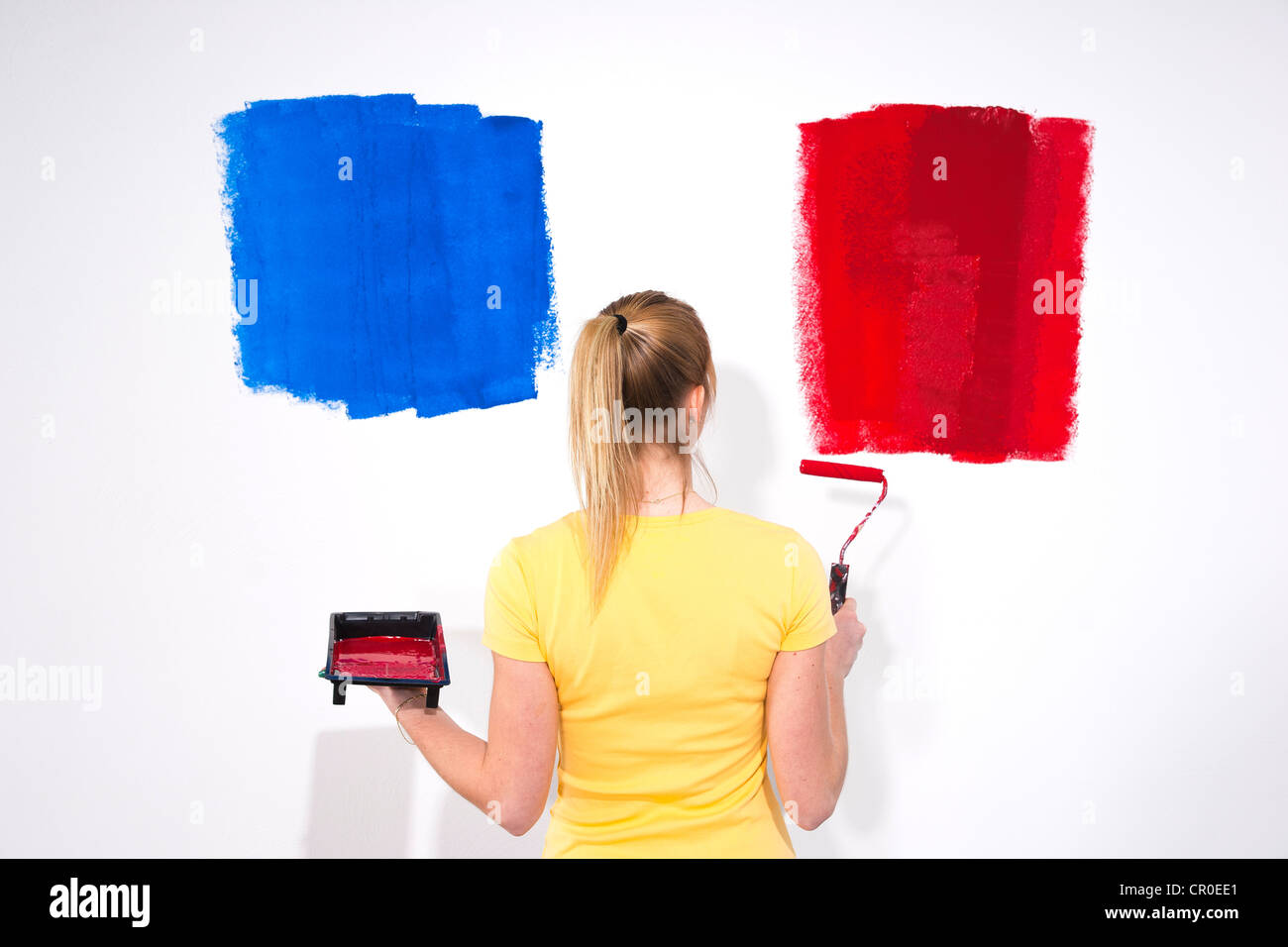 Giovane donna in piedi con un vassoio di vernice e un rullo di vernice davanti a una parete con campioni di vernice Foto Stock
