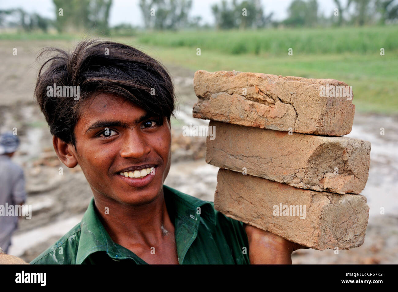 Sorridente giovane uomo che porta tre mattoni in una mano, sito in costruzione di un canale di irrigazione, Basti Lehar Walla village, Punjab Foto Stock