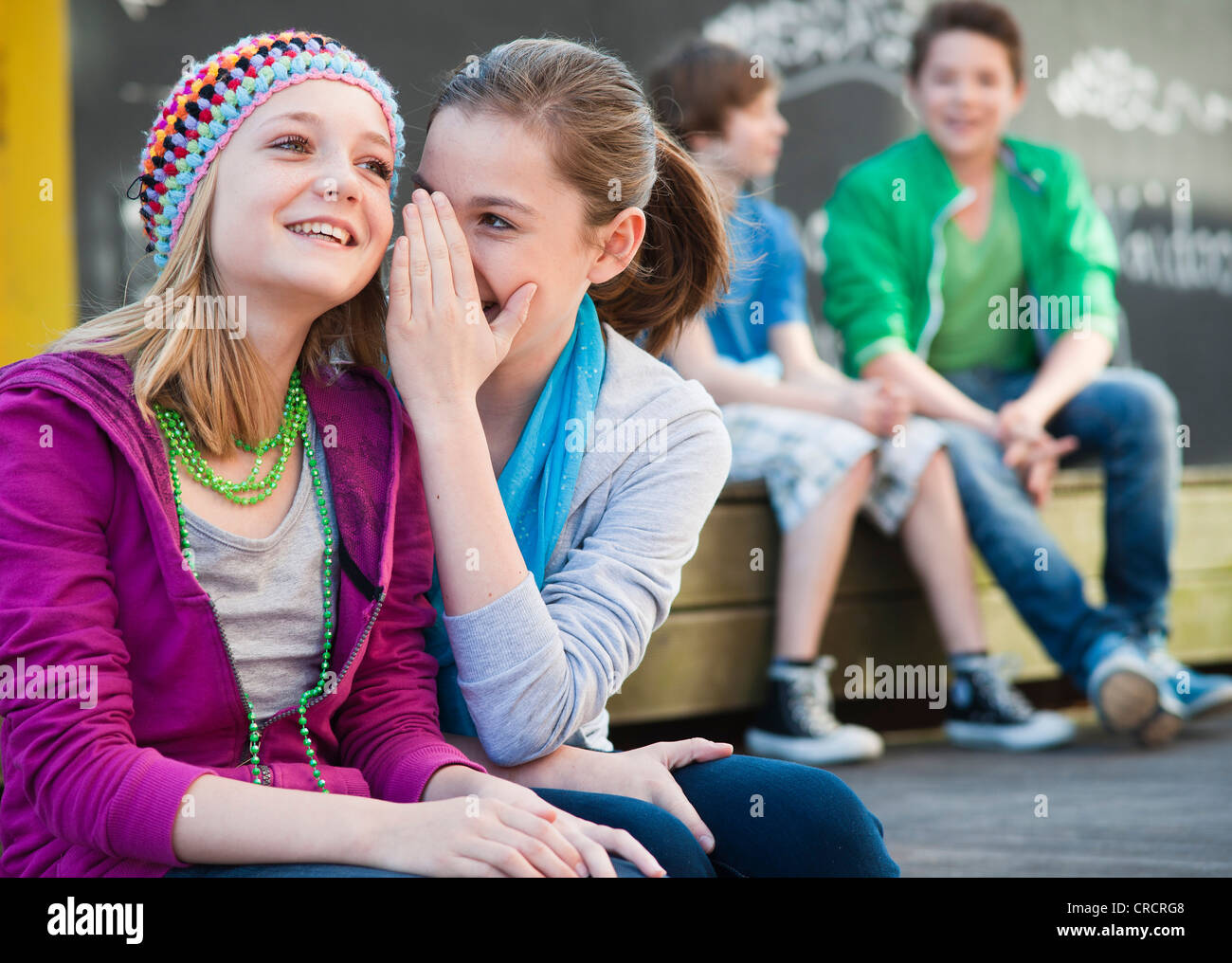 Due ragazze adolescenti whispering con due ragazzi in background Foto Stock