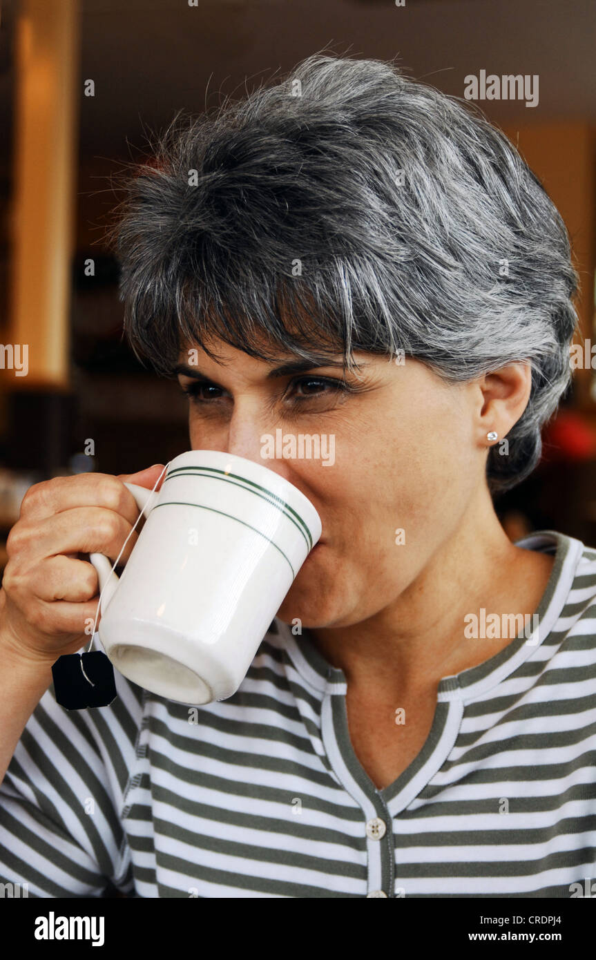 Una donna di mezza età di bere una tazza di tè mentre guardando oltre l'orlo del bicchiere e sorridente Foto Stock