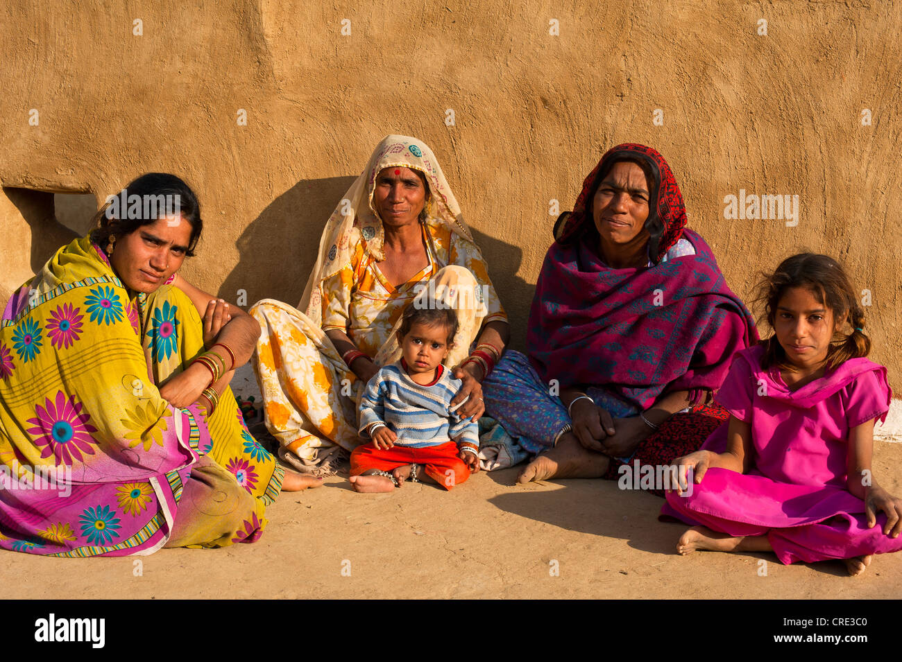 Molte donne e bambini che indossano vestiti tradizionali seduti di fronte al muro di una casa, il Deserto di Thar, Rajasthan, India Foto Stock