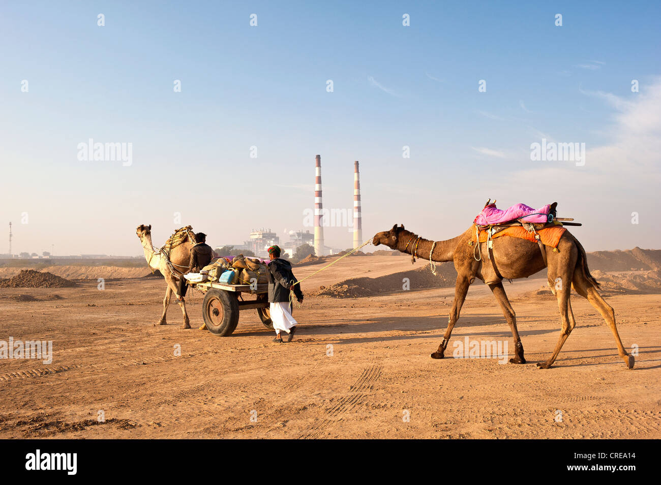 Due uomini indiano passando un impianto industriale con due enormi camini con i loro cammelli e un cammello carrello, il Deserto di Thar, Rajasthan Foto Stock