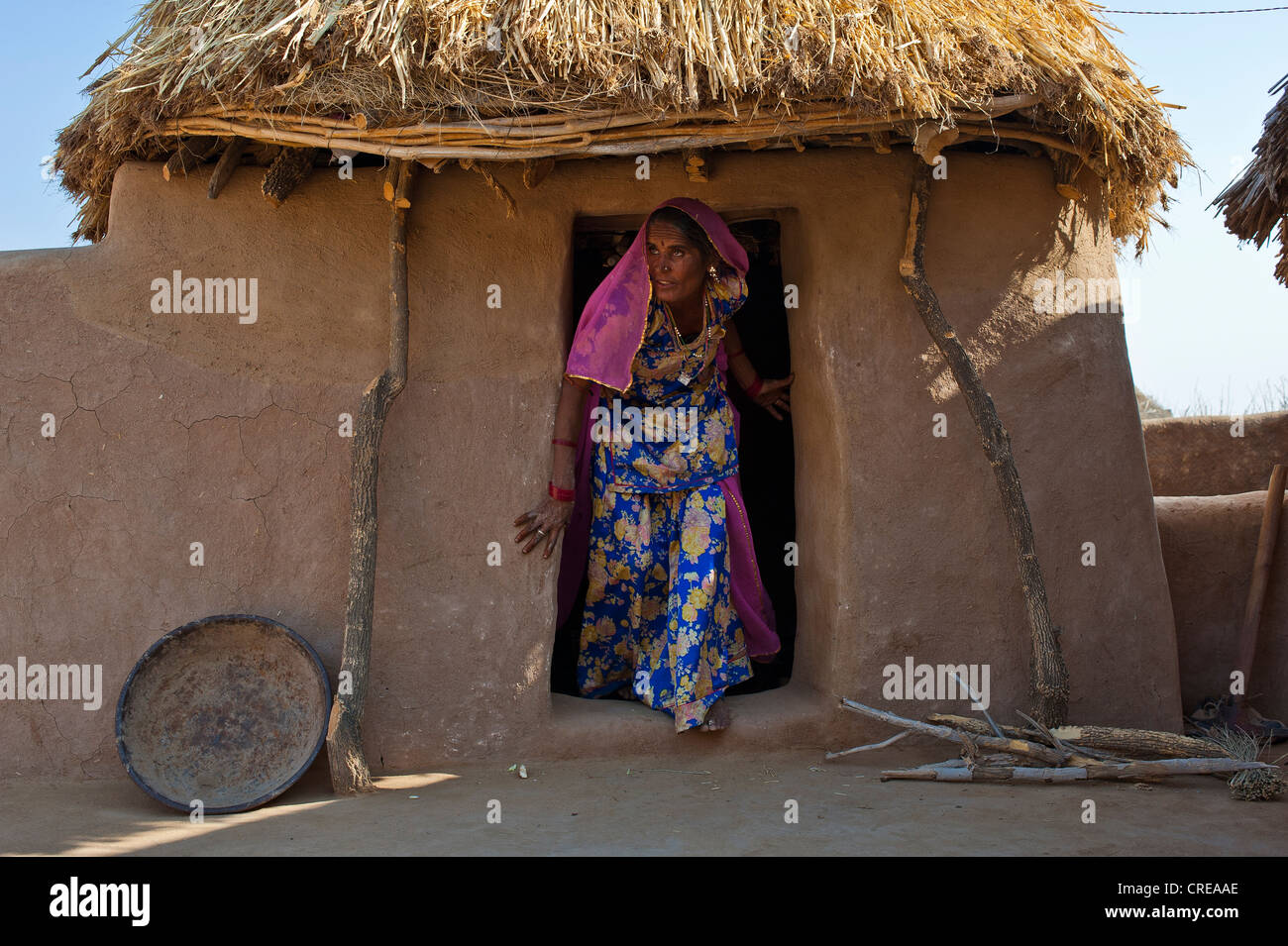 Donna indiana indossando un sari uscire dall'ingresso di una capanna, stovewood giace di fronte al rifugio, il Deserto di Thar Foto Stock