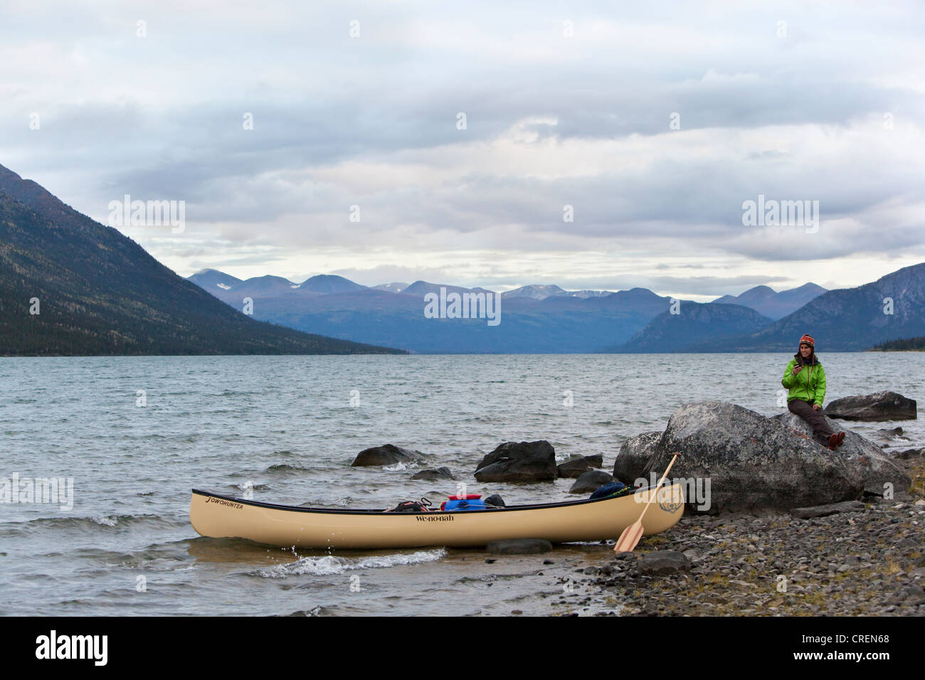 Giovane donna seduta su una roccia, canoa e pagaia in legno sulla riva, Kusawa lago, le montagne alle spalle, Yukon Territory, Canada Foto Stock