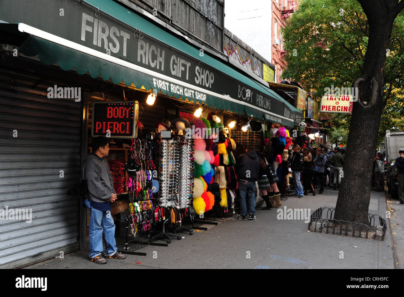 Oriental uomo in piedi al di sotto di neon rosso body piercing segno accanto al primo ricco Regali, St Mark, East Village, New York Foto Stock