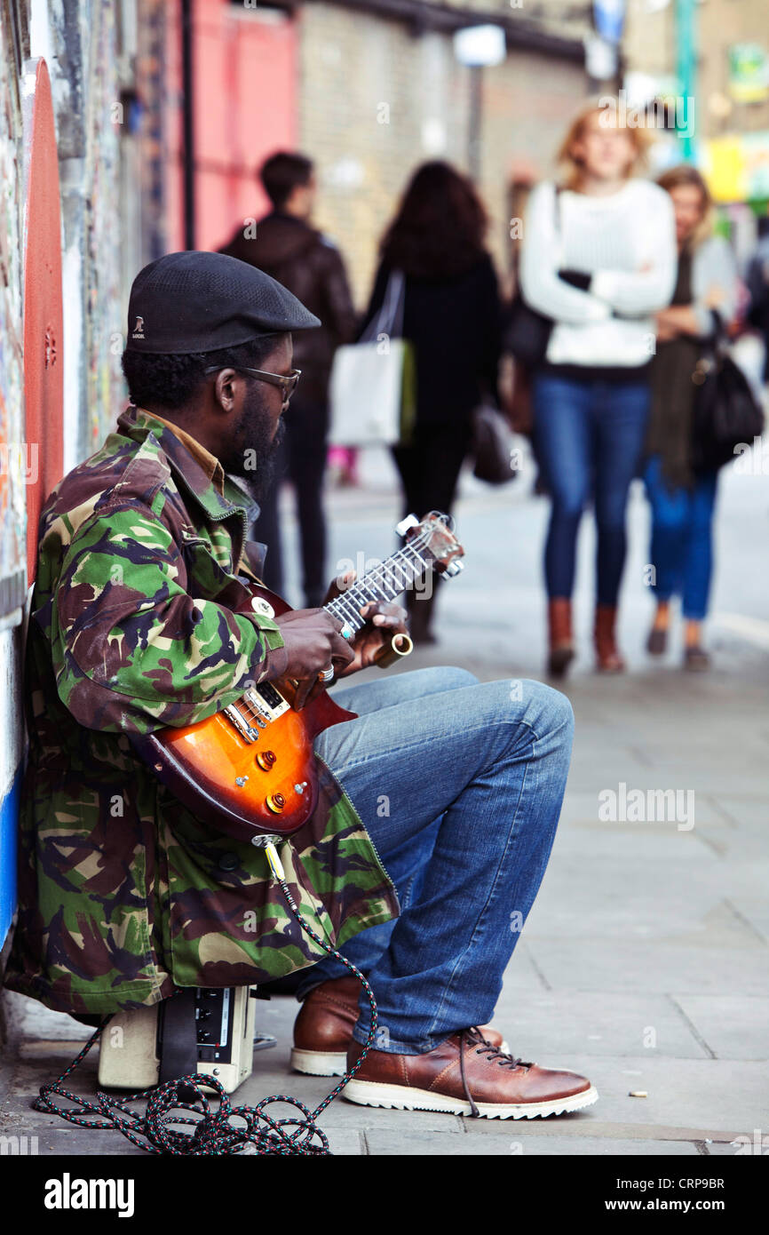 Ritratto completo di un busker maschio seduto sul marciapiede, suonando una chitarra elettrica, Brick Lane, Londra, Inghilterra, Regno Unito. Foto Stock