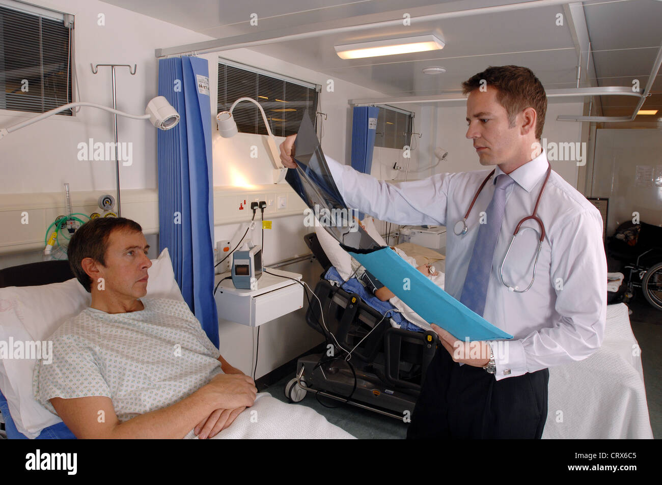 Un medico sulla sua ward round recensioni un x-ray durante un ospedale di consultazione al posto letto con la sua paziente di sesso maschile. Foto Stock