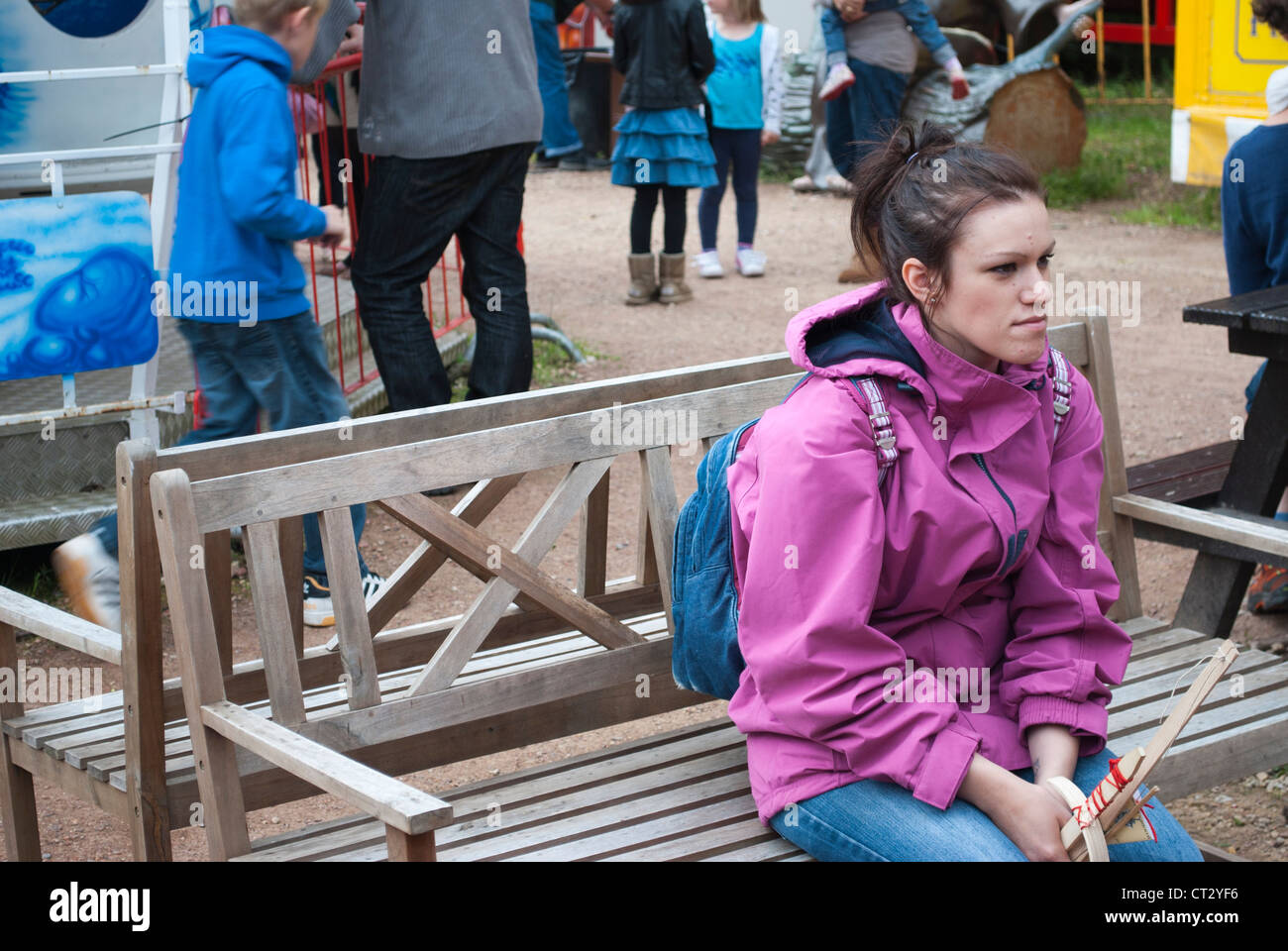 Giovani donne con capelli tirati indietro indossando rosa top con cappuccio e zaino blu sat su banco al fair con persone dietro Foto Stock