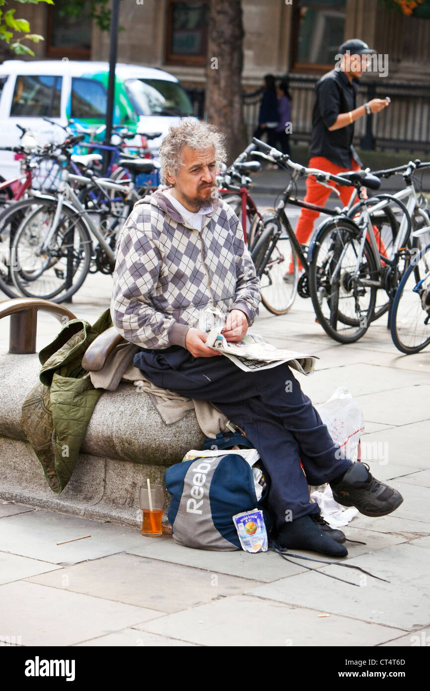 A piena lunghezza Ritratto di un senzatetto uomo seduto e tiene un giornale di Londra, Inghilterra, Regno Unito Foto Stock