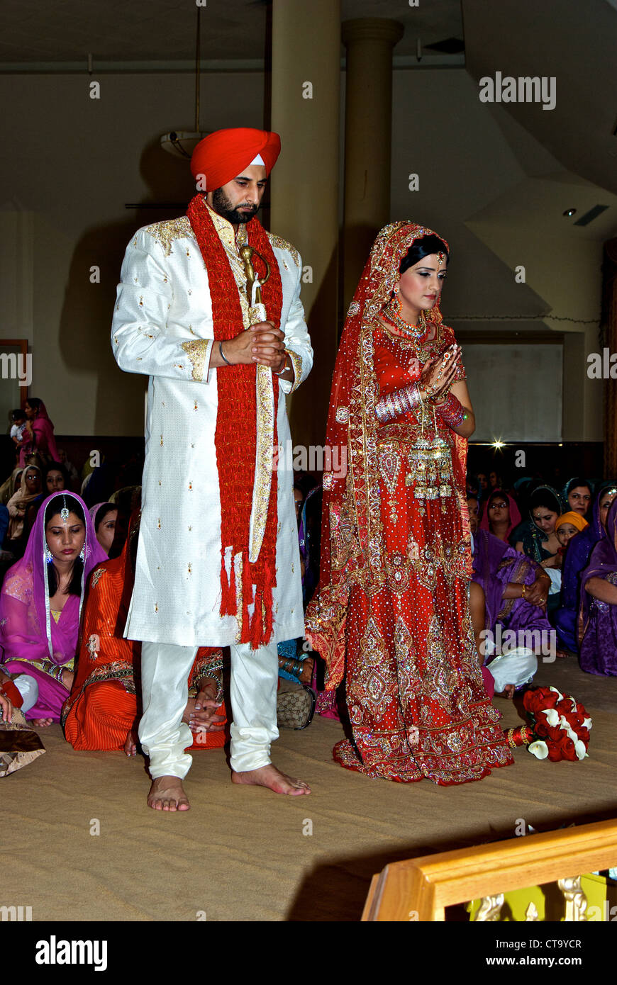 Est tradizionale indiano matrimonio Sikh groom luminoso turbante rosso vesti cerimoniali spada sposa rosso sari ricezione di preghiera di benedizione Foto Stock