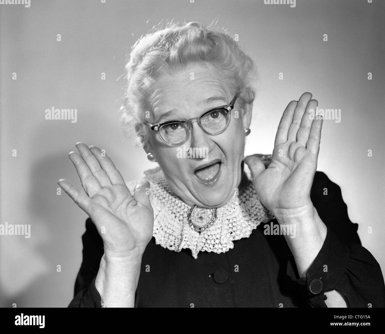 Anni sessanta donna anziana IN GRANNY bicchieri tenendo la mano fino in prossimità di faccia con la bocca aperta guardando la fotocamera Foto Stock