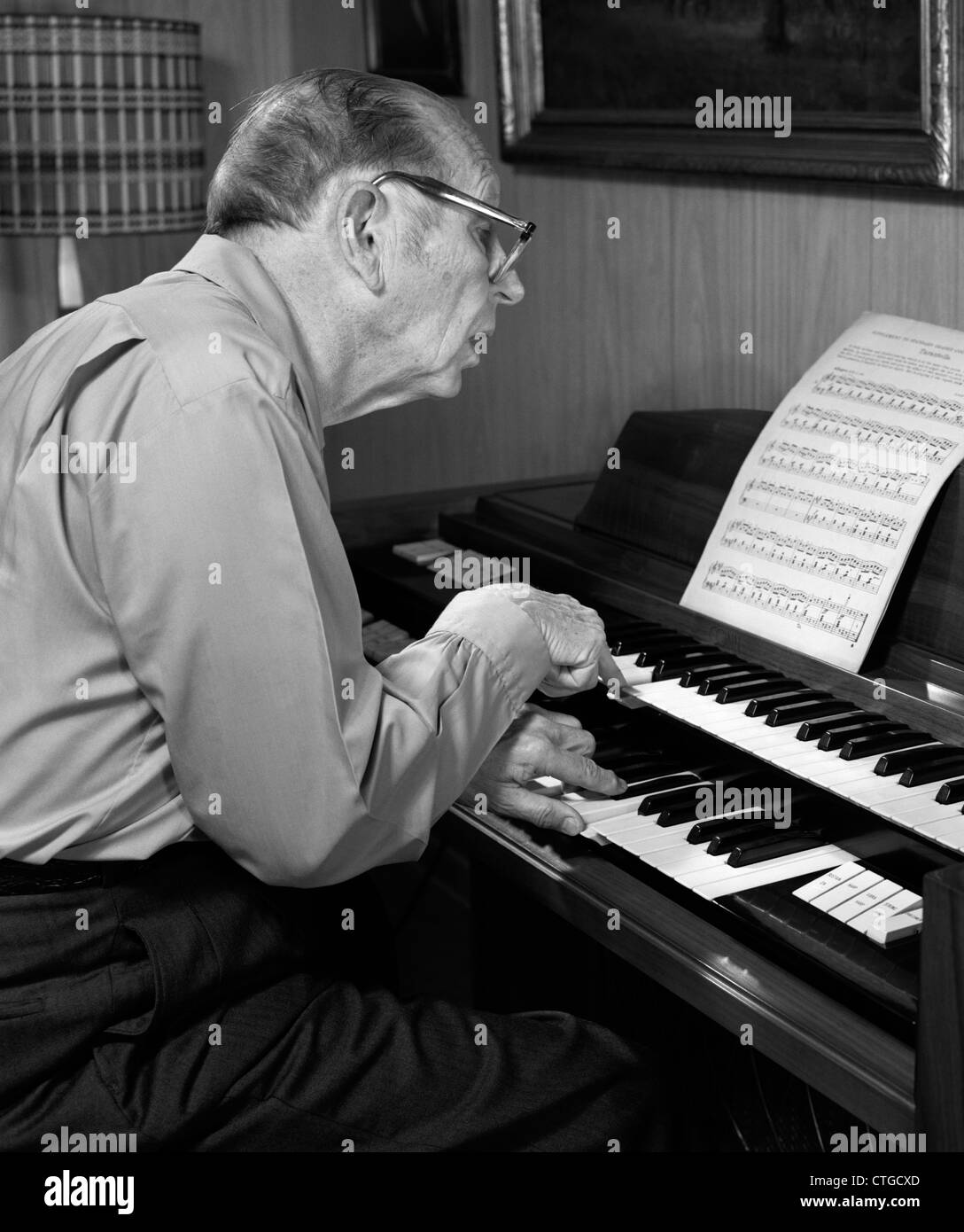 Anni Settanta uomo anziano CON GLI OCCHIALI INCLINATI IN AVANTI per leggere gli spartiti musicali durante la riproduzione di un organo Foto Stock
