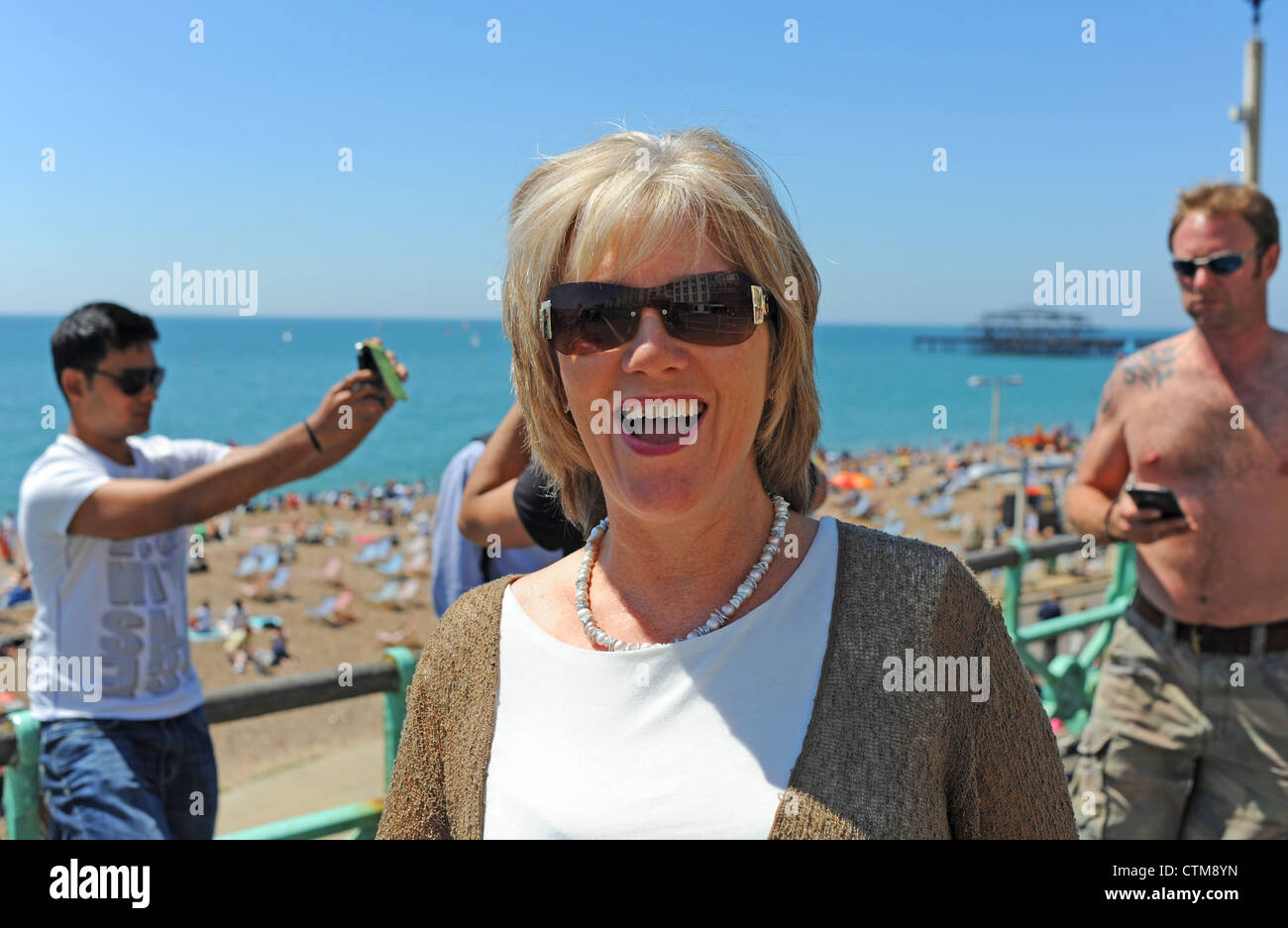 Mezza età attraente bionda femmina ridendo alla macchina fotografica sul lungomare di Brighton come due uomini dietro scattare fotografie UK estate Foto Stock