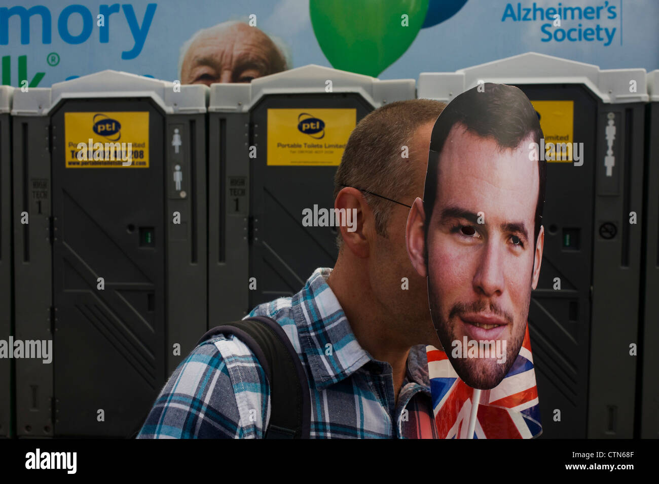 Con una carità billboard dietro, uno spettatore indossa una maschera con la faccia del Team GB ciclismo su strada hero Mark Cavendish nella prima giornata di gara del London 2012 Olympic 250km mens' la gara su strada. A partire da Londra centrale e passando la capitale più famosi luoghi di interesse prima di uscire in Inghilterra rurale all'estenuante Box Hill nella contea del Surrey. Sud-ovest locali londinesi rivestito il percorso sperando per il britannico Cavendish favorito per la vittoria di Team GB prima medaglia ma che sono poi stati delusi quando il Kazakistan è Alexandre Vinokourov alla fine ha vinto l'oro. Foto Stock