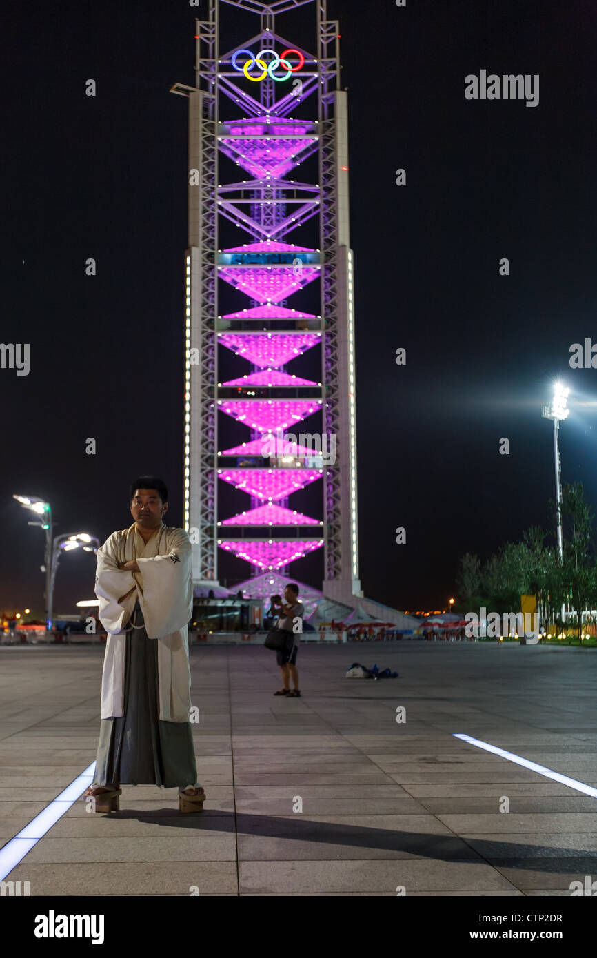 Pechino CINA - Agosto 16, 2008: uomo cinese in abito tradizionale pone di fronte alla torre olimpica con anelli illuminata di notte Foto Stock