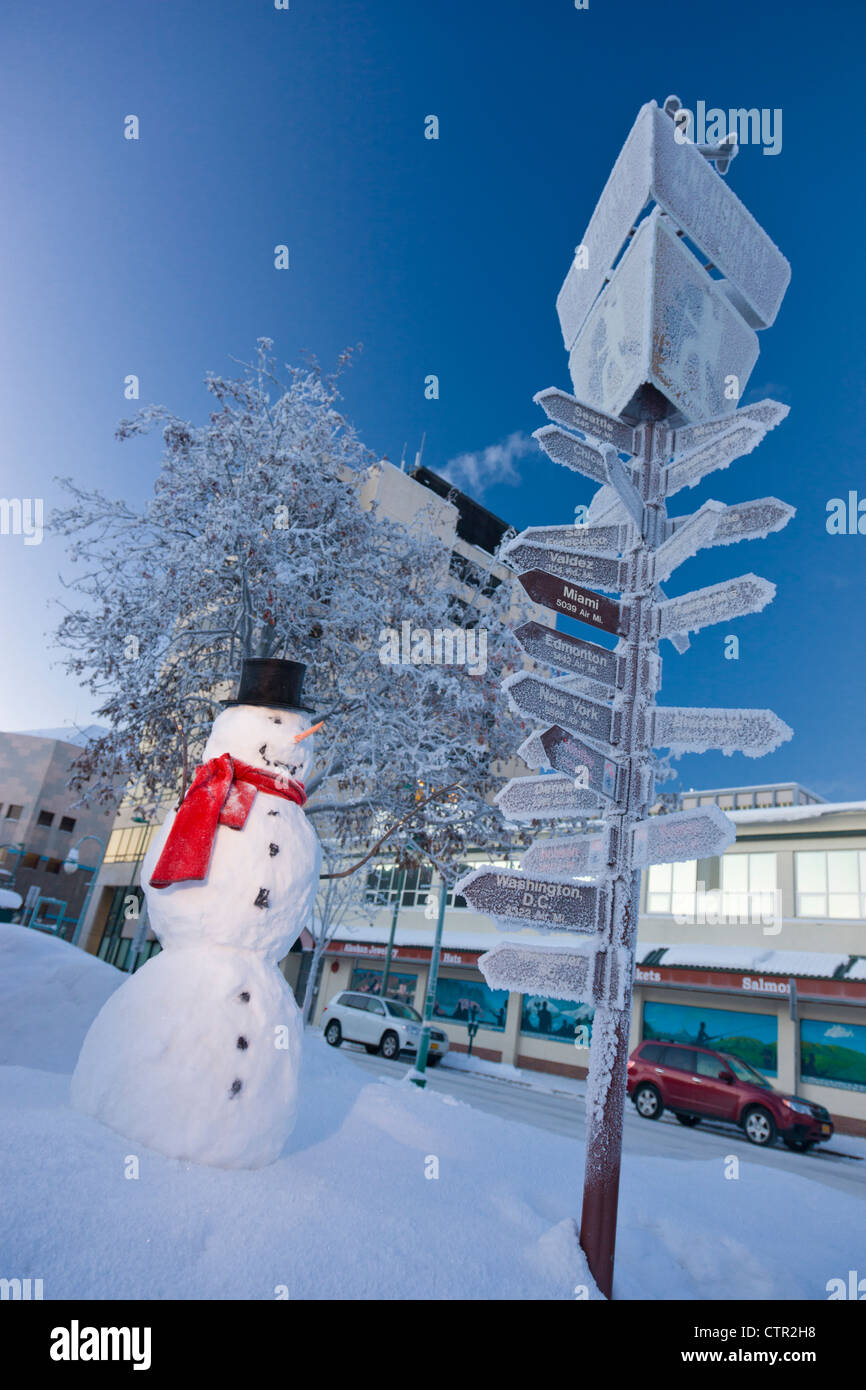 Pupazzo di neve vestita di nero top hat sciarpa rossa in piedi accanto chilometraggio da orientamento per i visitatori log cabin nel centro cittadino di Anchorage centromeridionale Foto Stock