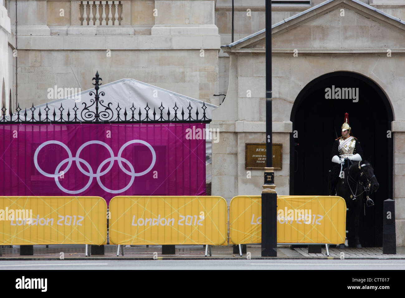 Anelli olimpici su un banner al di fuori delle Guardie a Cavallo in cui un soldato di cavalleria della famiglia siede immobile sul suo cavallo durante le Olimpiadi di Londra 2012. Ringhiere in ferro battuto sono visto dietro il banner presso l'impianto sportivo che ospita la pallavolo nel centro di Westminster dove gli uffici governativi sono situati. La casa britannica della cavalleria è classificata come un corpo a sé stante, e consiste di due reggimenti: vita delle guardie (esercito britannico) e il Blues e Royals (Royal Horse Guards e 1 ° dragoni). Essi sono il senior reggimenti regolari dell'esercito britannico, con tradizioni risalente al 1660. Foto Stock