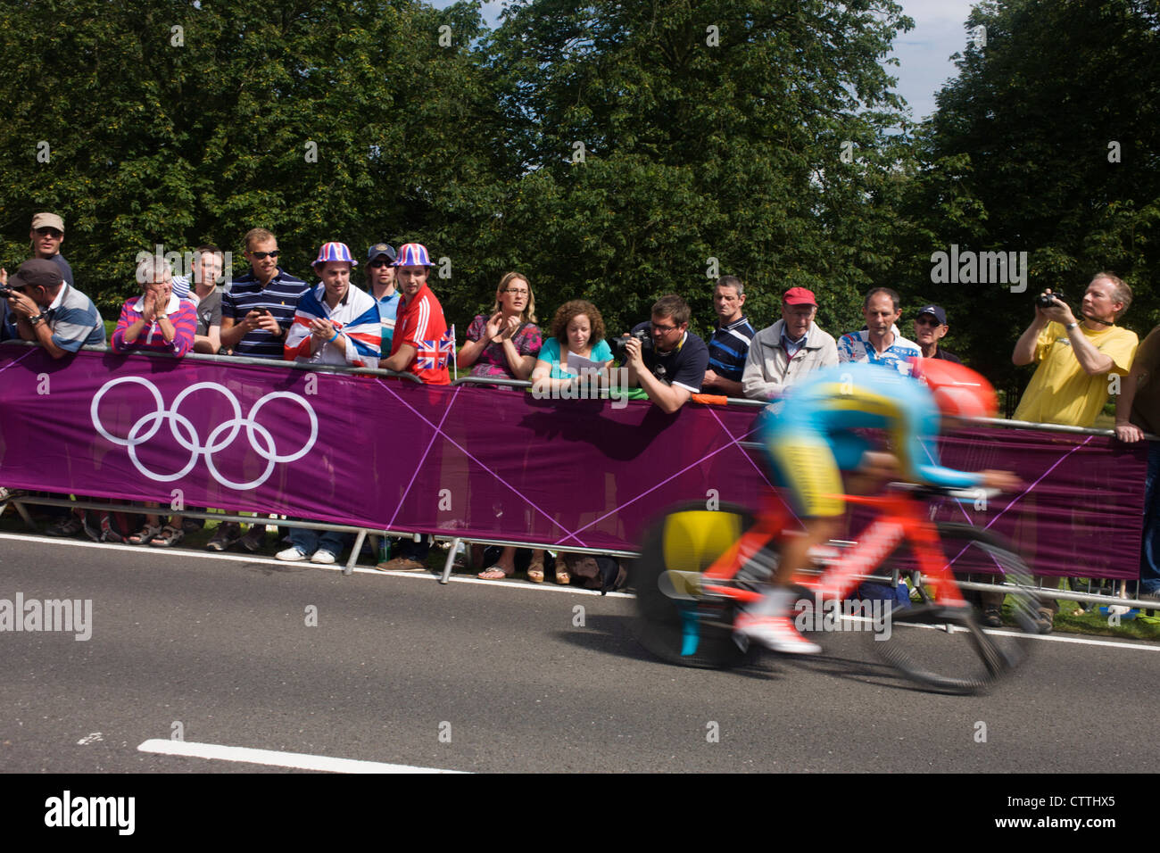 Un ciclista gare passato tifosi di rivestimento del percorso attraverso Bushy Park nel sud ovest di Londra, durante il London 2012 Olympic 44km maschile di ciclismo prova a tempo, infine vinto dalla squadra del GB Bradley Wiggins. Foto Stock