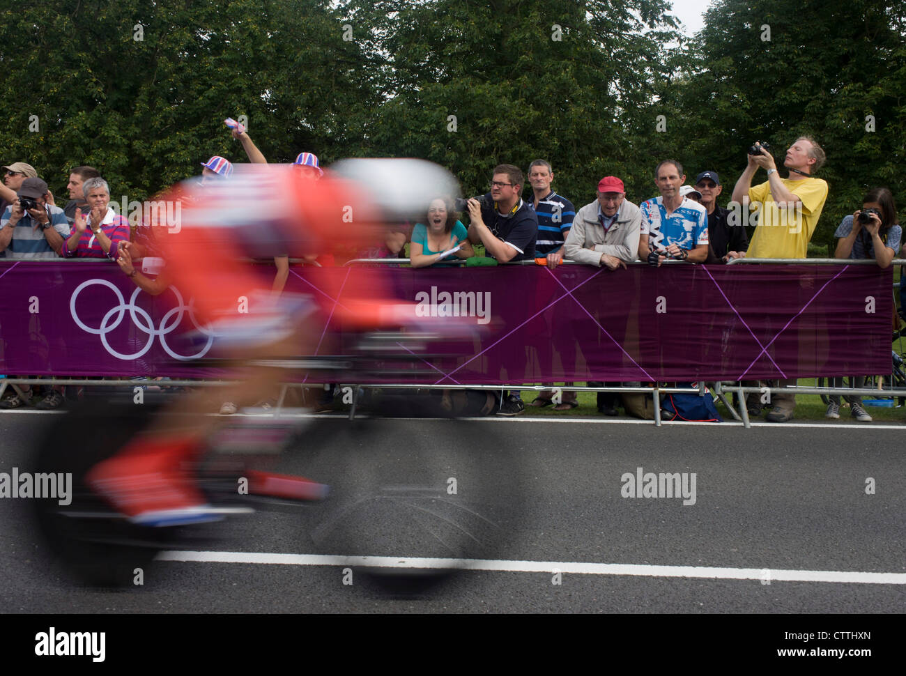 Un ciclista gare passato tifosi di rivestimento del percorso attraverso Bushy Park nel sud ovest di Londra, durante il London 2012 Olympic 44km maschile di ciclismo prova a tempo, infine vinto dalla squadra del GB Bradley Wiggins. Foto Stock