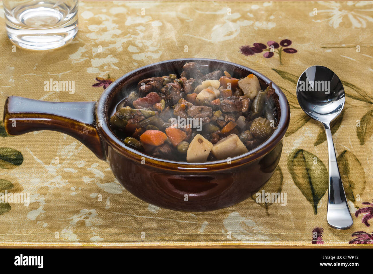 Recipiente di ceramica di carni bovine pot roast zuppa su tessuto di cartellone con cucchiaio e bicchiere di acqua Foto Stock