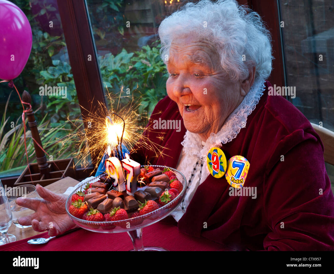 Torta di compleanno anziana signora scintilla divertimento sorridente celebrando il suo 97th compleanno a casa di famiglia con torta frizzante con 9-7 badge di compleanno e palloncini Foto Stock